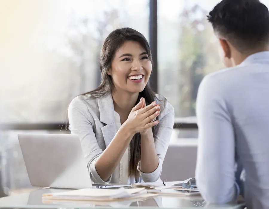 Professional meeting between a smiling business professional and a client seated across a desk with a laptop and documents, engaged in conversation in a bright office setting.