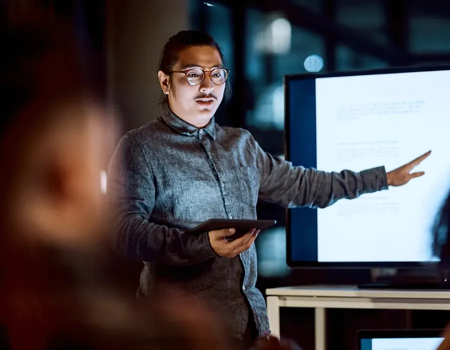IT project manager presenting information on a large screen while holding a tablet and speaking to colleagues in a modern office.