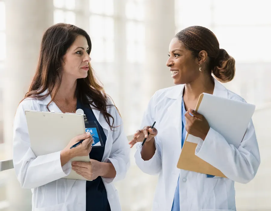 Two nurse practitioners in white lab coats stand in a bright hospital corridor, talking and smiling at each other while holding clipboards and pens.