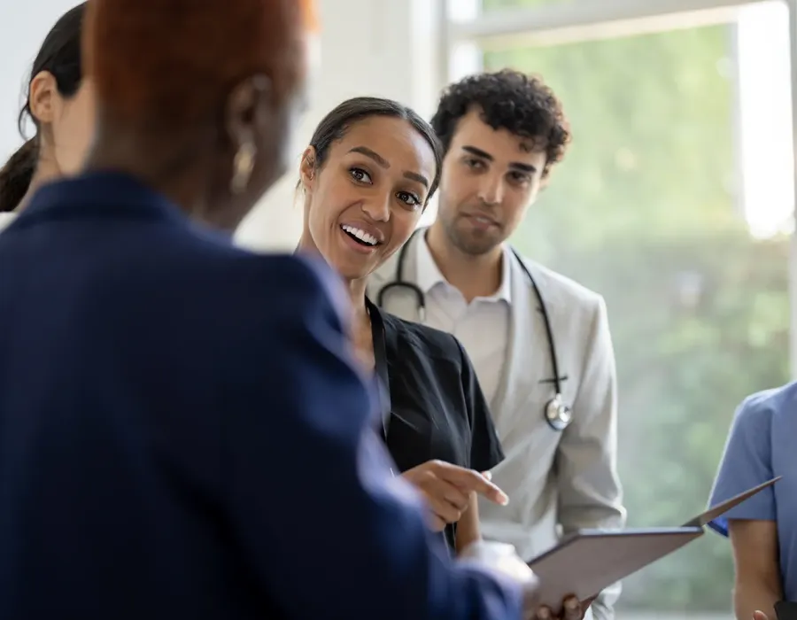 Smiling nurse holding a clipboard while speaking with a healthcare team during a patient care discussion.