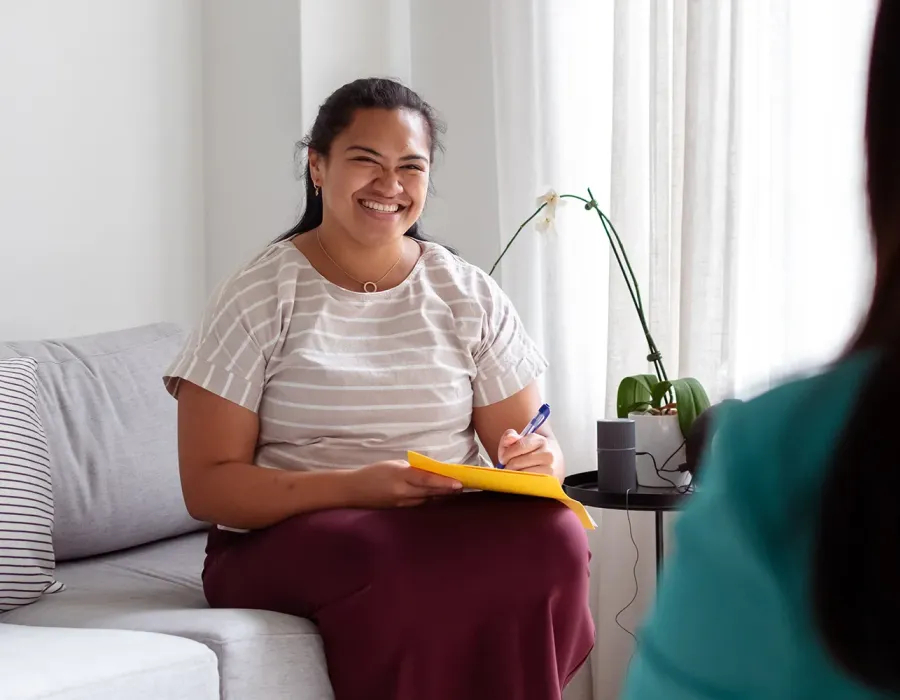 A smiling social worker and former RN sits on a couch holding a notepad and pen during a conversation with another person in a bright, comfortable room.