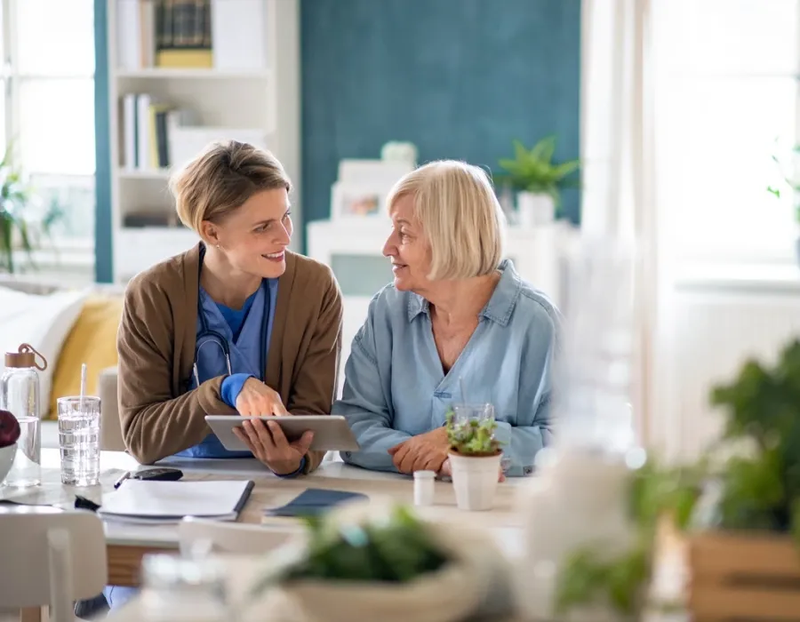 A holistic nurse in blue scrubs with a stethoscope sits beside an older adult, smiling and reviewing information together on a tablet at a table in a bright home or care setting.