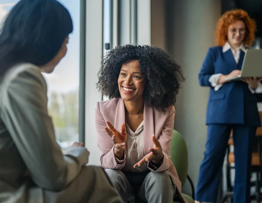 Three behavioral health professionals converse in a modern office, with one woman smiling and gesturing while another listens and a third works on a laptop in the background.