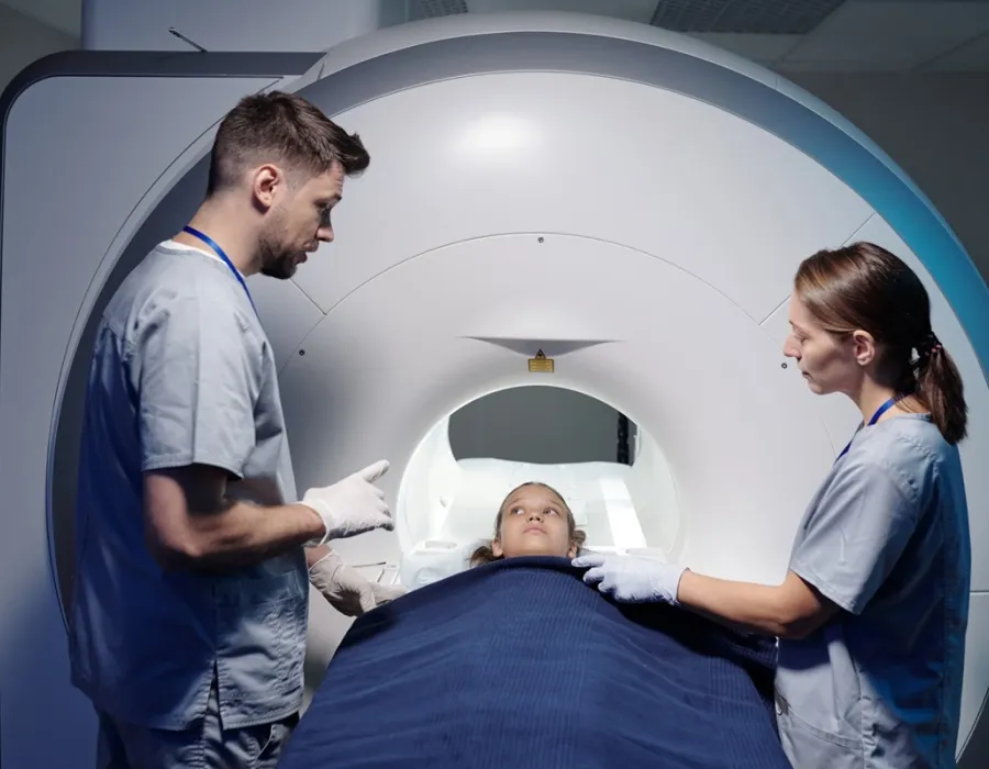An MRI Tech and Imaging Assistant stand beside a patient lying on an MRI scanner table, preparing the patient for imaging.