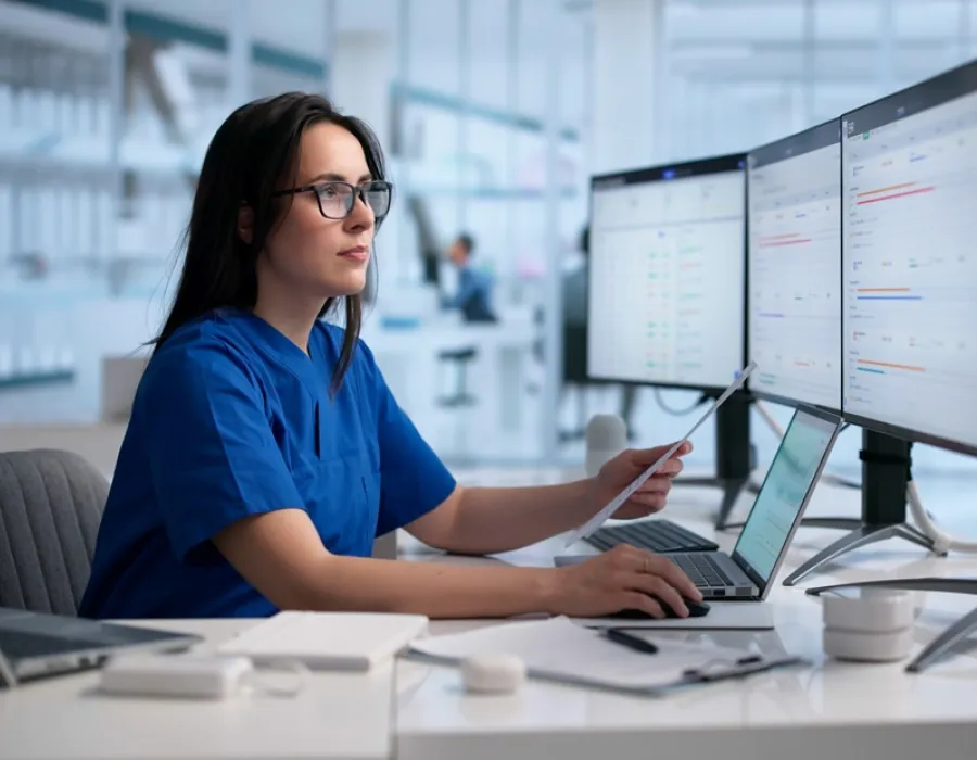 A nurse sitting by her computer at a desk. She's typing on a separate laptop while holding a document in her left hand.