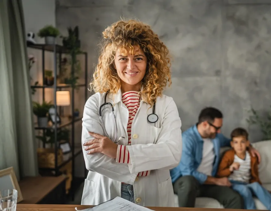 Smiling FNP in a white coat and stethoscope standing with arms crossed while an adult and child sit together in the background.
