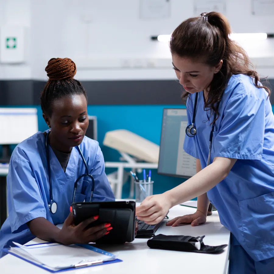 Two medical assistants in blue scrubs viewing patient charts on tablet