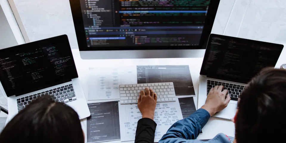 Two computer programmers seated at desk in front of multiple monitors showing coding software