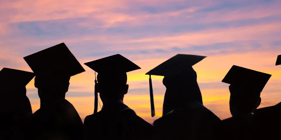A group of seven nursing graduate students look of into the distance as the sun sets. They are wearing caps and gowns.