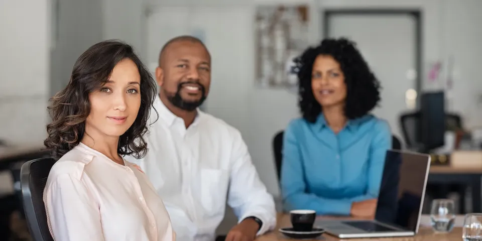 a group of three business professionals are setting in a board room. They are looking at the camera. 