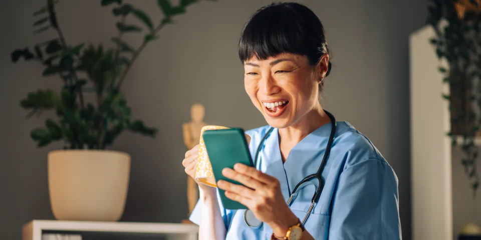 Asian female nurse in blue scrubs laughs heartily while looking at her mobile phone, holding a mug of coffee. The background features potted plants, bookshelves, and a small wooden figure.