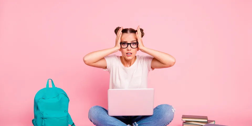 Full legs, body, size portrait of confused girl in white T-shirt and blue jeans sitting on floor with laptop on knees isolated on bright pink background.