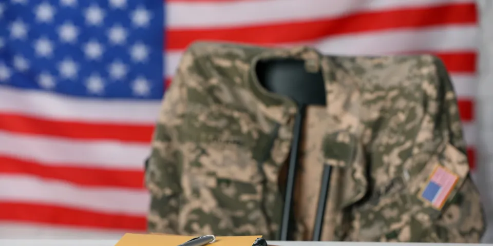 Notebooks and pen on white wooden table, chair with soldier uniform against flag of United States indoors. Military education
