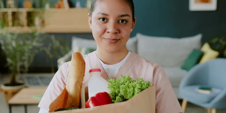 Young nurse holding paper bag full of healthy food while looking at camera and smiling widely