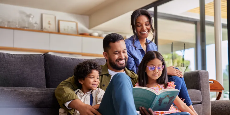 Mixed race father reading a story book to daughter and son while sitting with beautiful wife on couch. Young multiethnic man sitting on floor carpet reading a fairy tale to his kids. Happy parents holding book and spending time with children on a happy weekend.