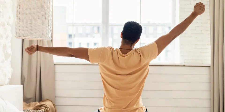 Arabic Man Sitting On Bed Back To Camera And Stretching Arms Awaking After Good Sleep In Modern Bedroom At Home, Looking Out Of Window. Comfort And Coziness. Good Morning Concept