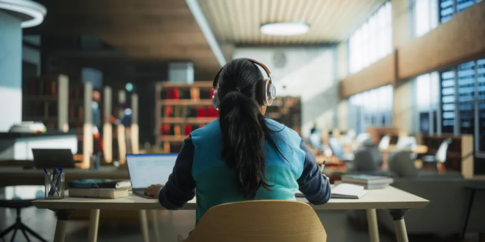 Female Student Wearing Headphones while Working on University Homework in a Public Library. Woman Sitting Behind a Desk, Using Laptop Computer and Writing Down Notes in Notebook. Footage from the Back