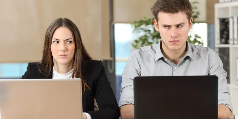 Front view of two angry businesspeople using computers disputing at workplace and looking sideways each other with anger