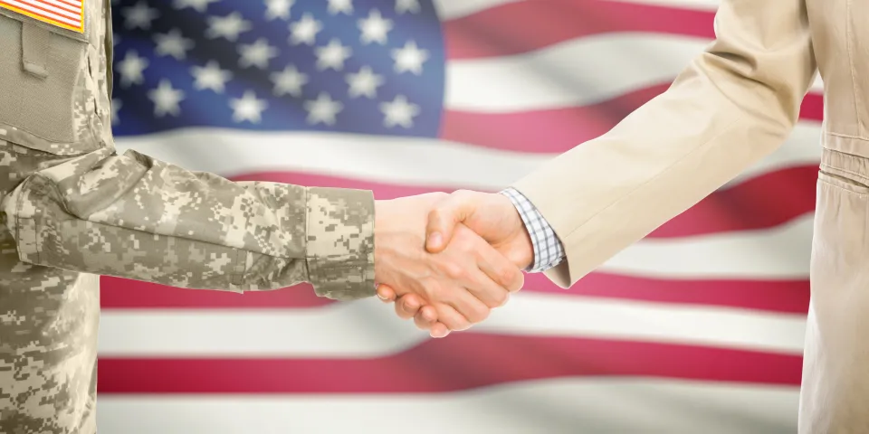 American soldier in uniform and civil man in suit shaking hands with national flag on background - United States