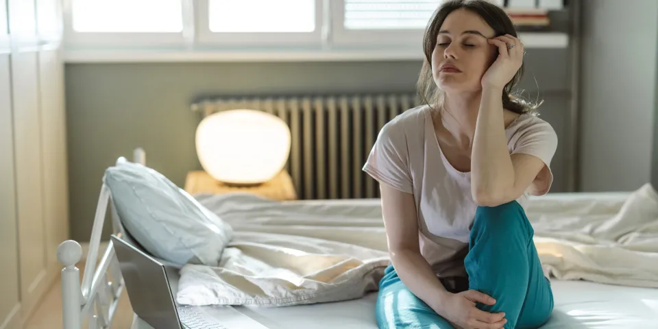 A young woman sitting on her bed in a sunny bedroom, appearing deep in thought and showing signs of tiredness. The scene suggests contemplation, relaxation, or a morning routine in a cozy setting.