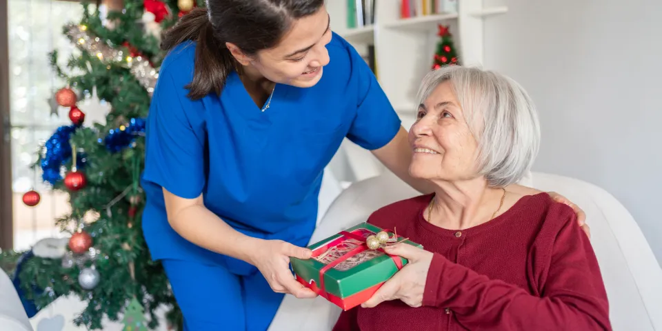 Home Caregiver Hugging And Giving Presents Senior Woman For Christmas At Nursing Home