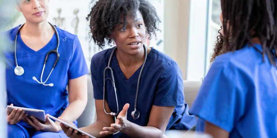 Nurses in blue scrubs sit together in a team meeting, discussing patient care while holding tablets.