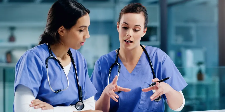 Two nurses in blue scrubs discuss treatment notes at a desk with a tablet and clipboard.