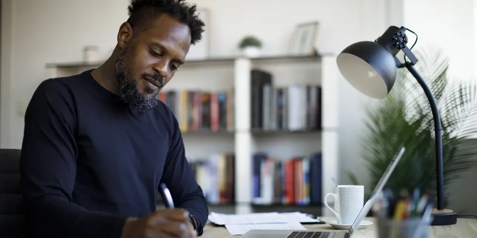 A student writes in a notebook at a desk with a laptop, phone, lamp, and bookshelves nearby.