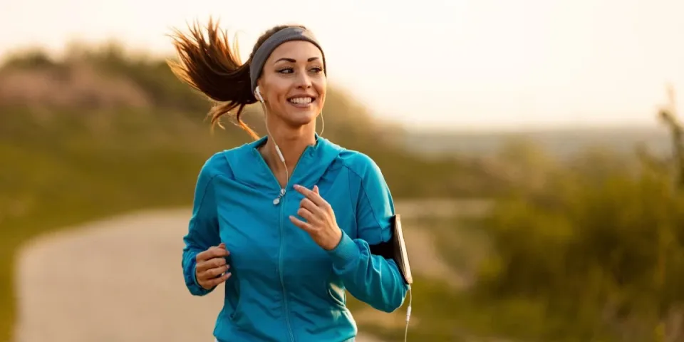 Woman smiling while jogging outdoors on a winding path, wearing earbuds and a blue jacket with a phone armband.