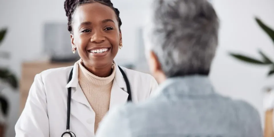 A smiling nurse practitioner wearing a white coat and stethoscope speaks with an older patient during an appointment.