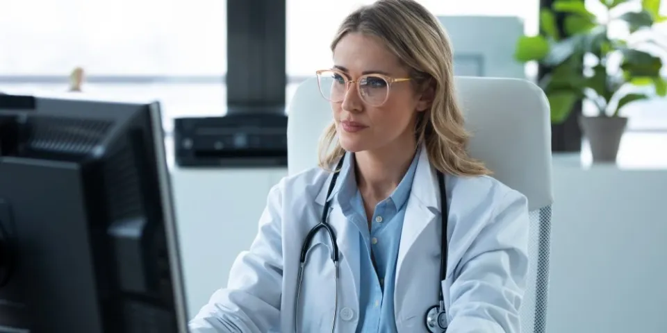 A nurse practitioner wearing a white lab coat and stethoscope sits at a desk using a computer in a bright medical office.