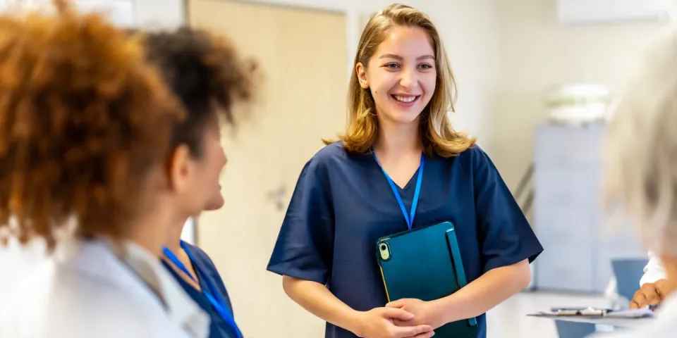 Smiling nursing student in scrubs holding a tablet while speaking with healthcare professionals in a clinical setting.