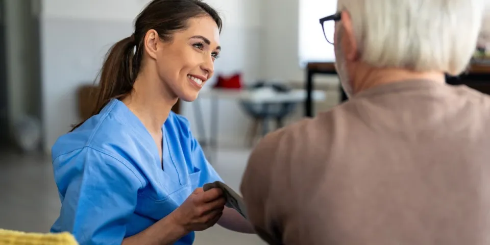 Smiling home health nurse in blue scrubs checks an older adult’s blood pressure during a patient care visit.