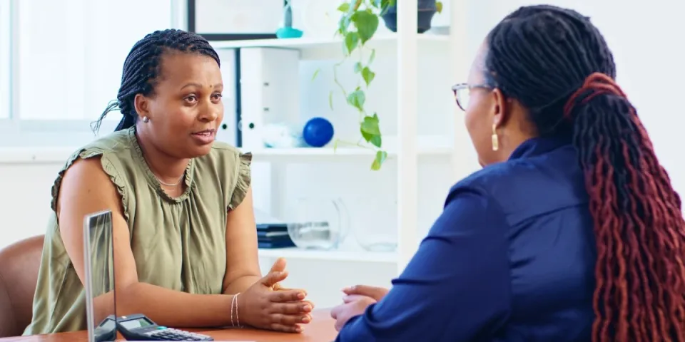 A human services worker speaking with a client in an office, engaged in a supportive one-on-one conversation with a laptop and office shelves in the background.