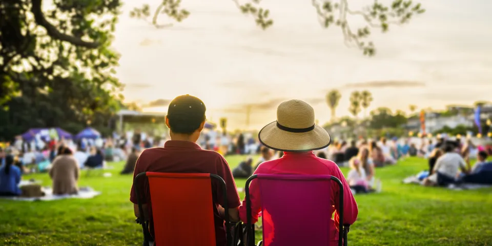 Couple sitting on the picnic chairs, listening to the music concert. Unrecognizable crowd of people in the park.