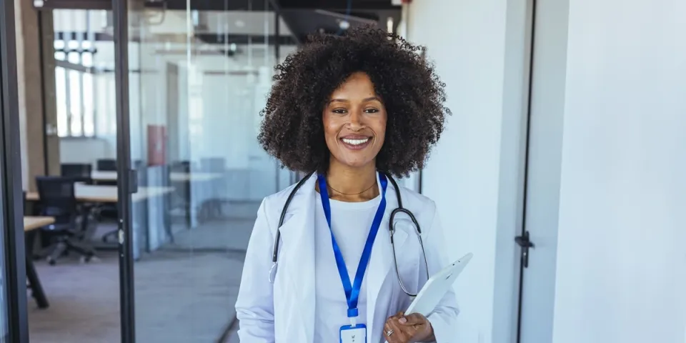 Smiling nurse practitioner in a white coat with a stethoscope holds a tablet in a modern medical office.