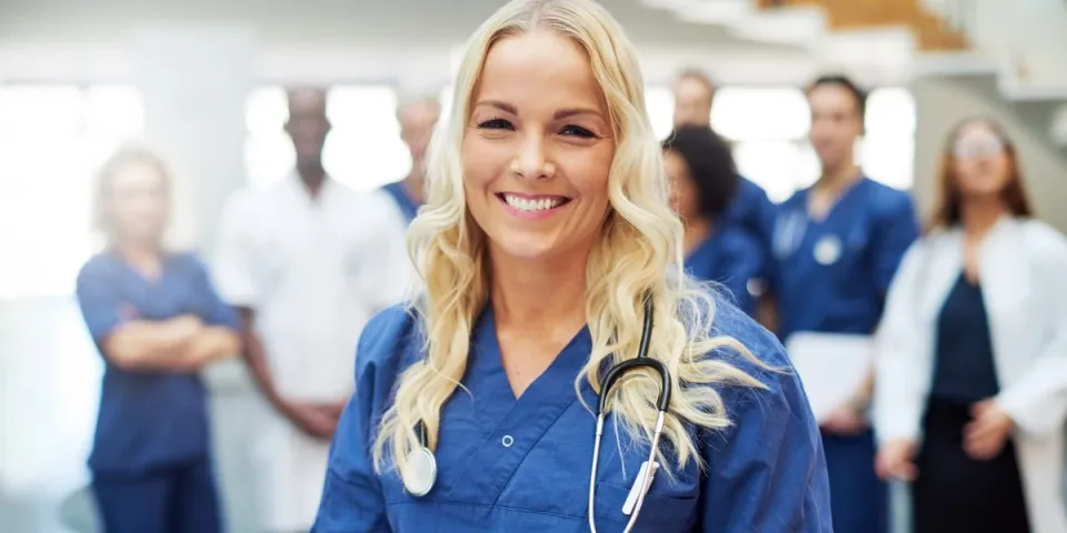 Smiling RN who is pursuing a BSN in blue scrubs with a stethoscope stands in the foreground while a multidisciplinary medical team gathers in the background of a healthcare facility.