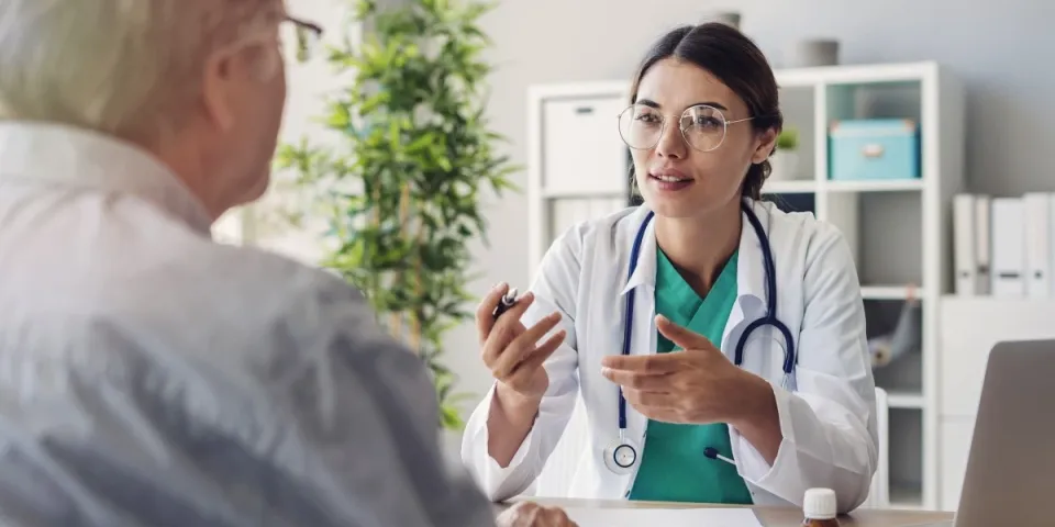 Nurse with a DNP wearing a stethoscope discusses care with an older patient during an office visit, seated across a desk with a laptop and paperwork.