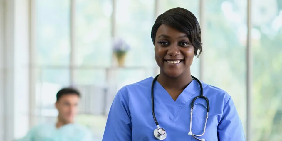 Smiling nurse in blue scrubs with a stethoscope and tablet standing in a bright hospital room, with a patient resting in a bed blurred in the background.