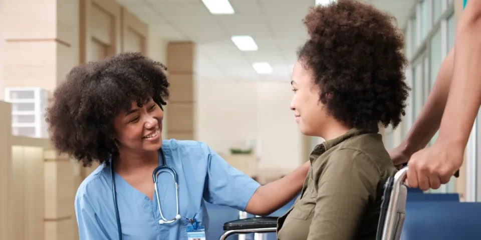 Registered nurse in blue scrubs speaks reassuringly with a patient seated in a wheelchair in a hospital hallway.