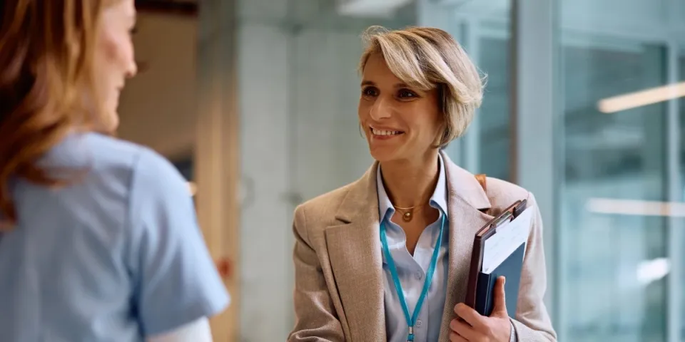 Professional shakes hands with a healthcare worker while holding a clipboard, suggesting a meeting or career opportunity in a healthcare setting.