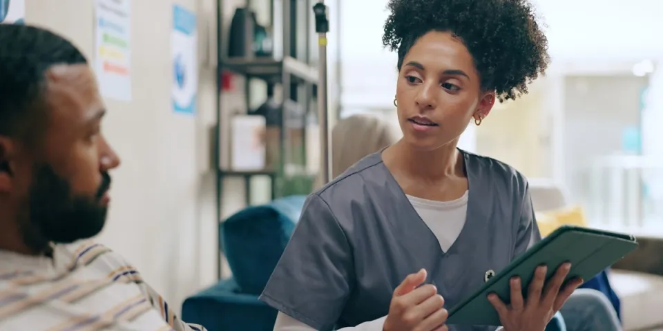 Registered nurse who graduated from an MSN Direct Entry program in scrubs discusses information on a tablet with a seated man in a home setting.