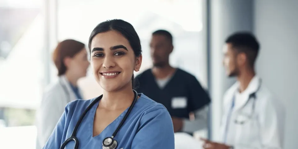 RN in blue scrubs with a stethoscope smiles confidently at the camera while colleagues collaborate in the background of a clinical setting.