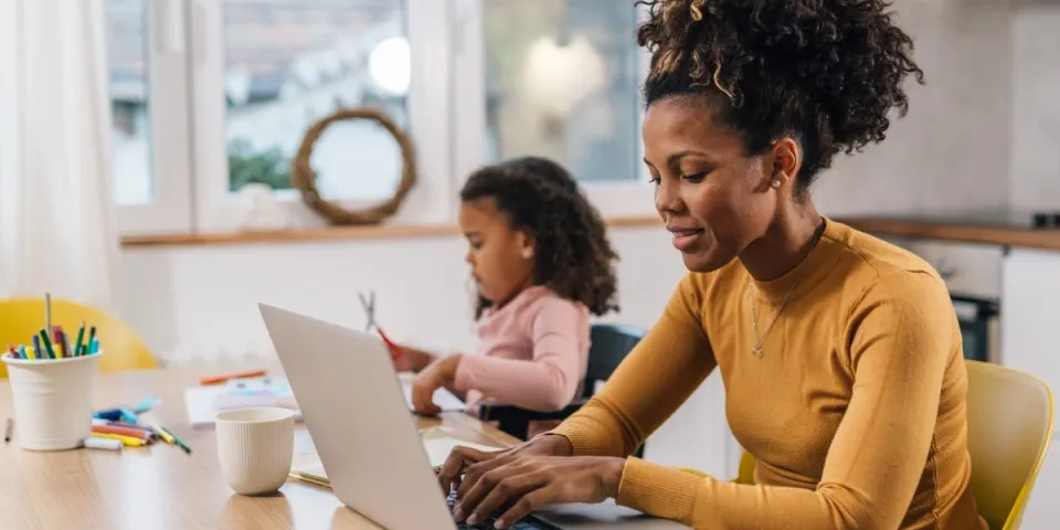 Parent works on a laptop at a kitchen table while a young child sits nearby doing arts and crafts, balancing work and family responsibilities at home.