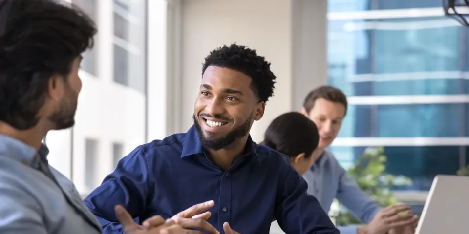 Four coworkers sit around a table in a bright office. A smiling man in a navy shirt talks with a colleague in the foreground while two others work near an open laptop in the background.