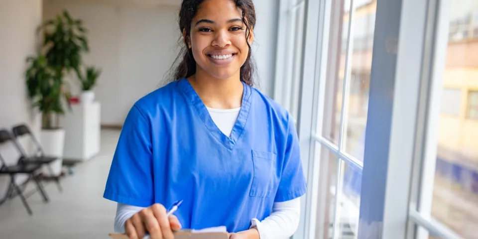 A smiling nurse in blue scrubs holds a clipboard and pen while standing beside large windows in a bright healthcare facility.