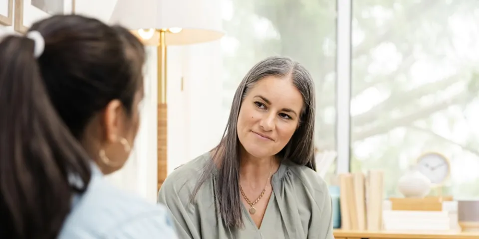 A holistic nurse listens attentively to a patient while holding a notepad and pen during a one-on-one conversation in a calm setting.