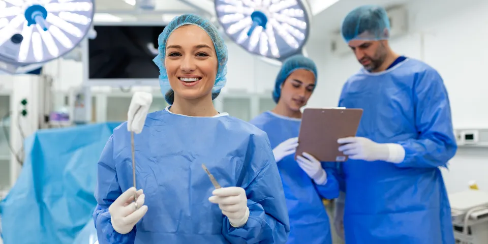 ortrait of Smiling Female Surgeon in Blue Scrubs Holding Surgical Scalpel and Medical Forceps in Modern Operating Room with Professional Medical Team in Background