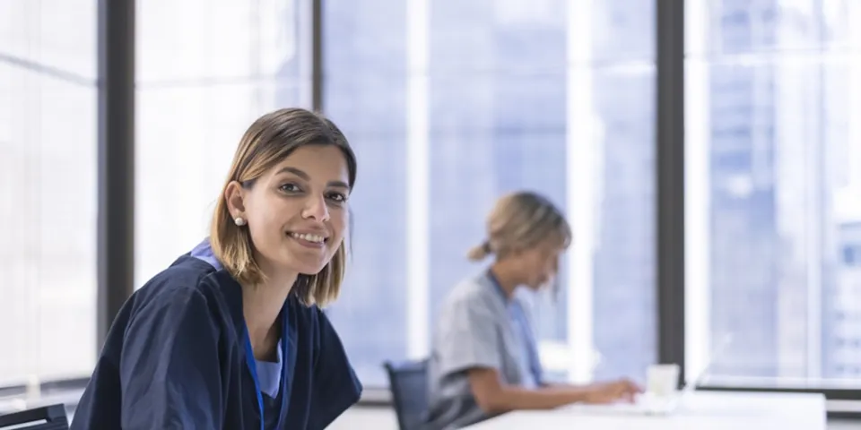 A nursing student studying at a table writes notes in a notebook while another student works quietly in the background near large windows.