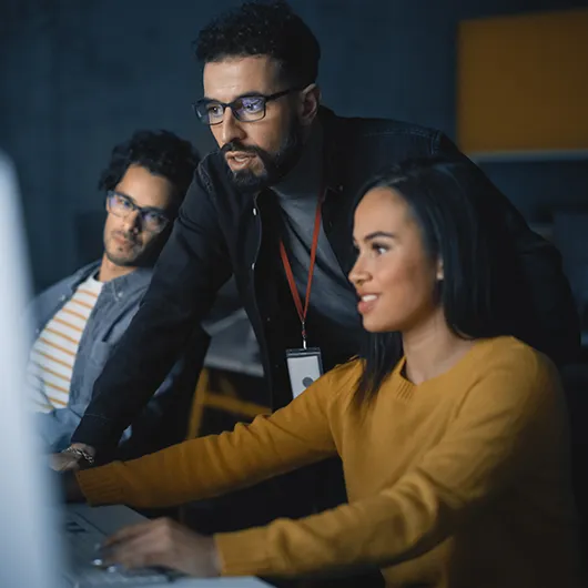 Three professionals collaborate in a modern office setting, focused on a computer screen; a woman in a yellow sweater types while two male colleagues observe and discuss the project.